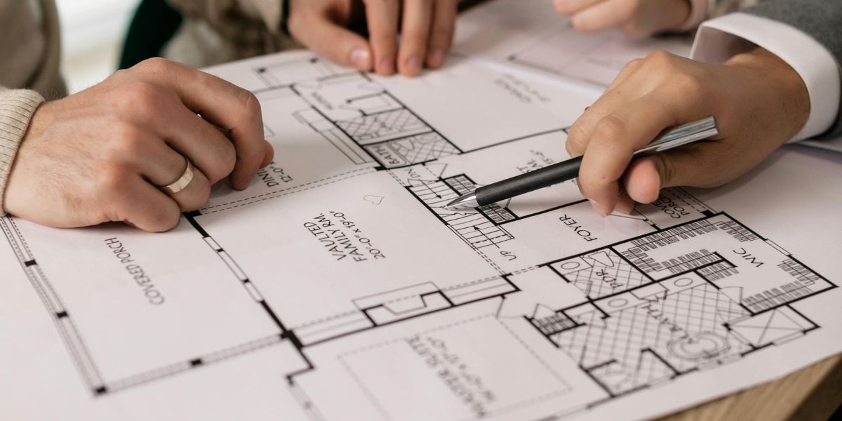 Close-up of hands pointing with a pen at a detailed floor plan or blueprint, indicating specific rooms and features.