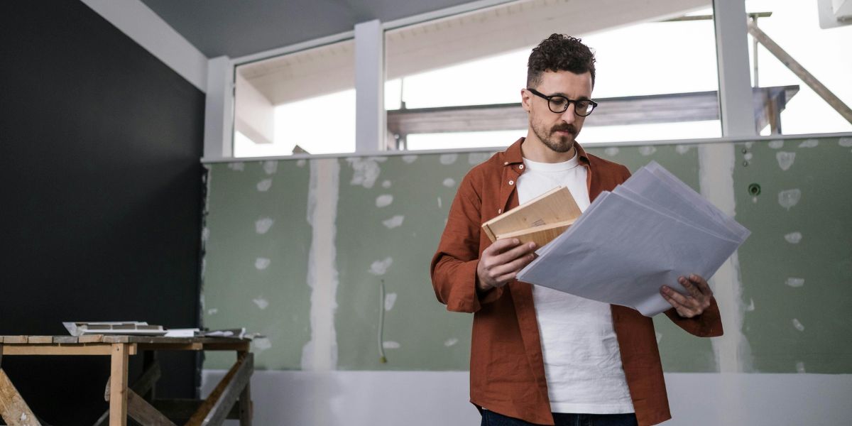 A man in glasses and a rust-colored shirt holds architectural plans and a wooden block, standing in a room under construction with green drywall and dark accent walls