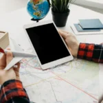 Person holding a miniature airplane model in one hand while using a tablet with a map and globe on the table, planning a travel journey.