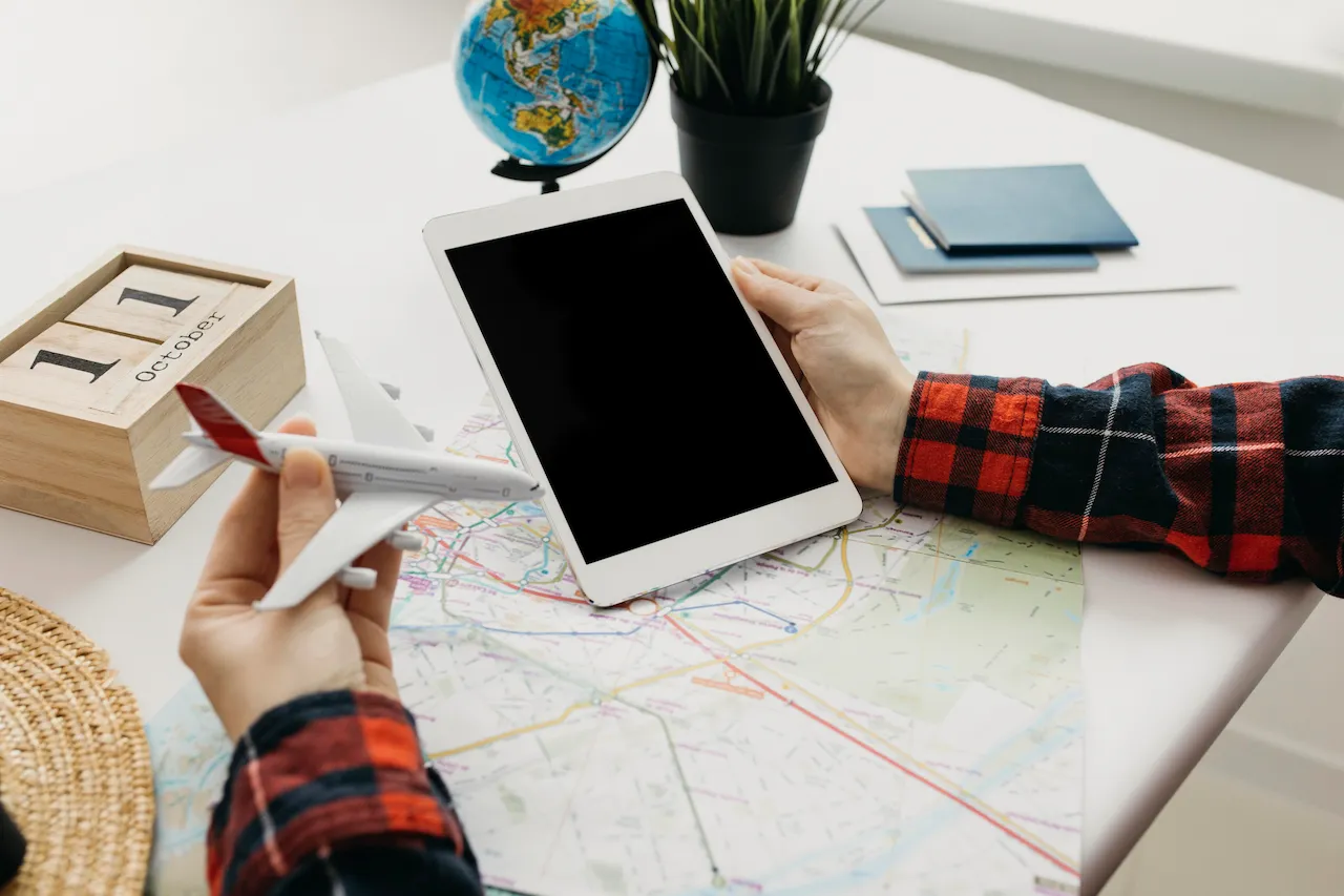 Person holding a miniature airplane model in one hand while using a tablet with a map and globe on the table, planning a travel journey.