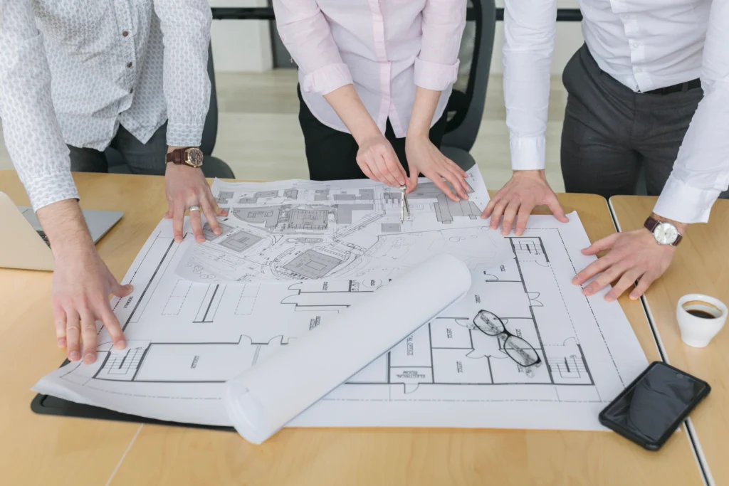 Professional team of architects and engineers reviewing building floor plans and site blueprints on a large office desk.