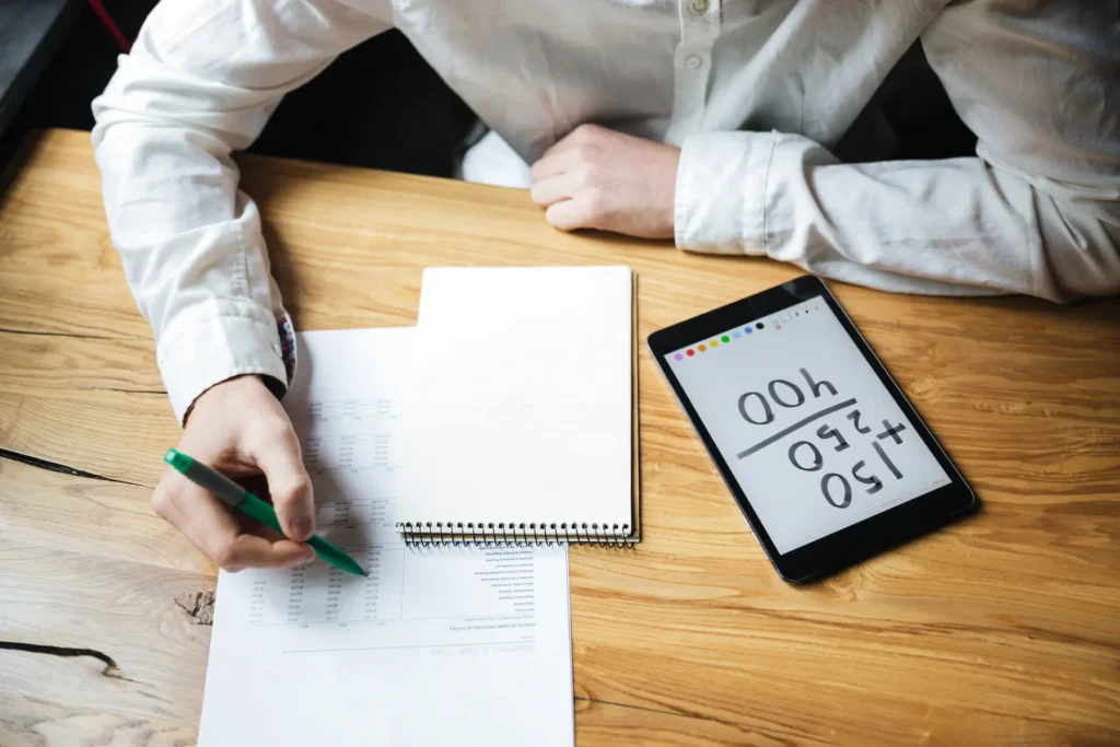 Person reviewing financial documents with a calculator on a tablet and taking notes at a wooden desk.