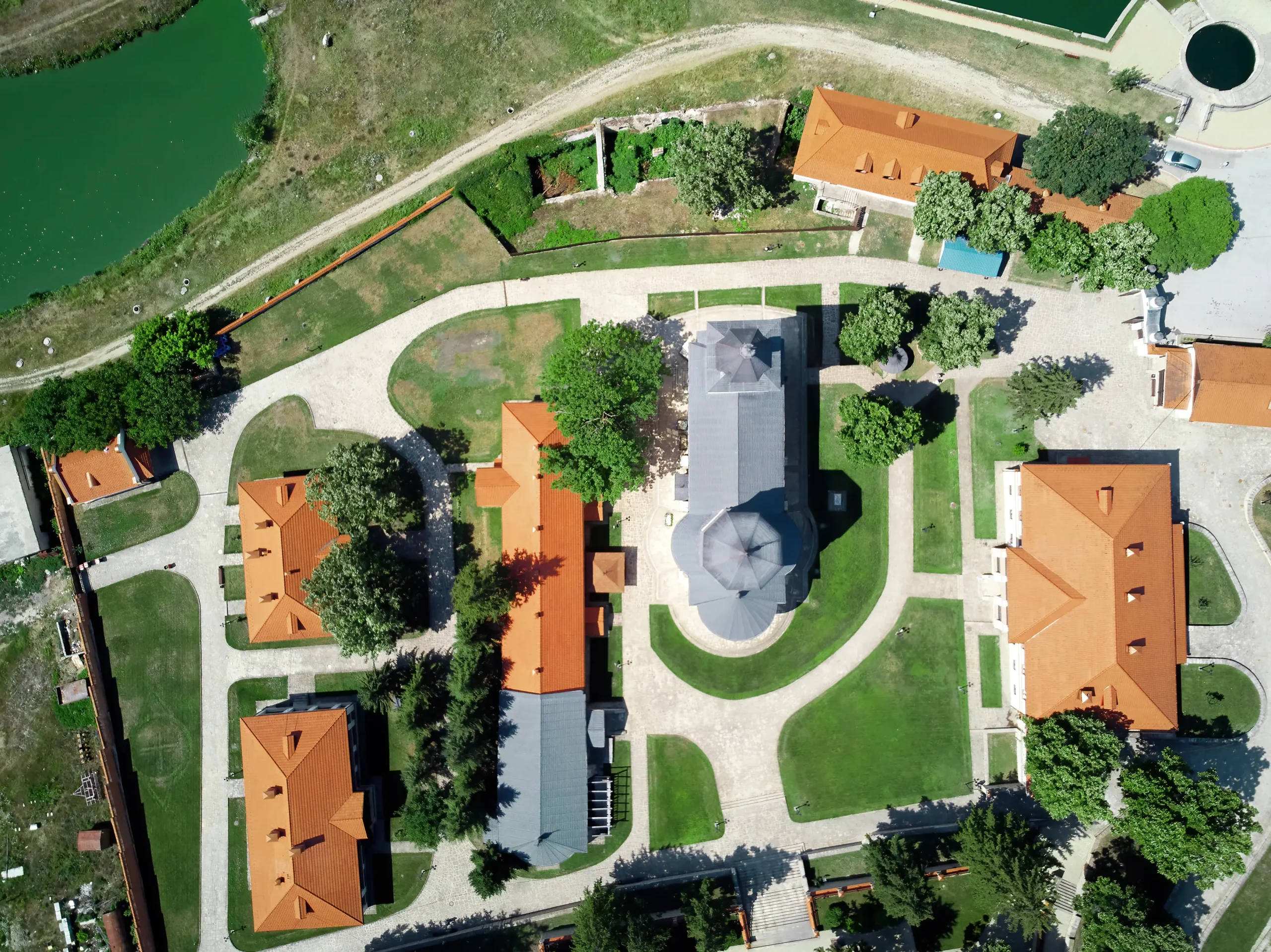 Aerial top-down view of a landscaped courtyard complex with historic buildings, red-tiled roofs, curved pathways, green lawns, and a central church-like structure near a water body.