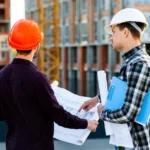 Construction engineer and architect wearing hard hats reviewing blueprints together at a residential building construction site with multi-story structure in the background.