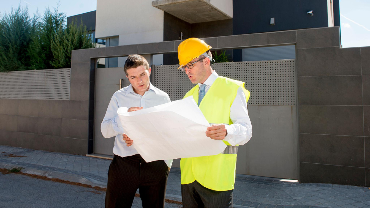 An architect in a yellow hard hat and safety vest reviewing blueprints with a client or developer outside a modern two-story residential house.