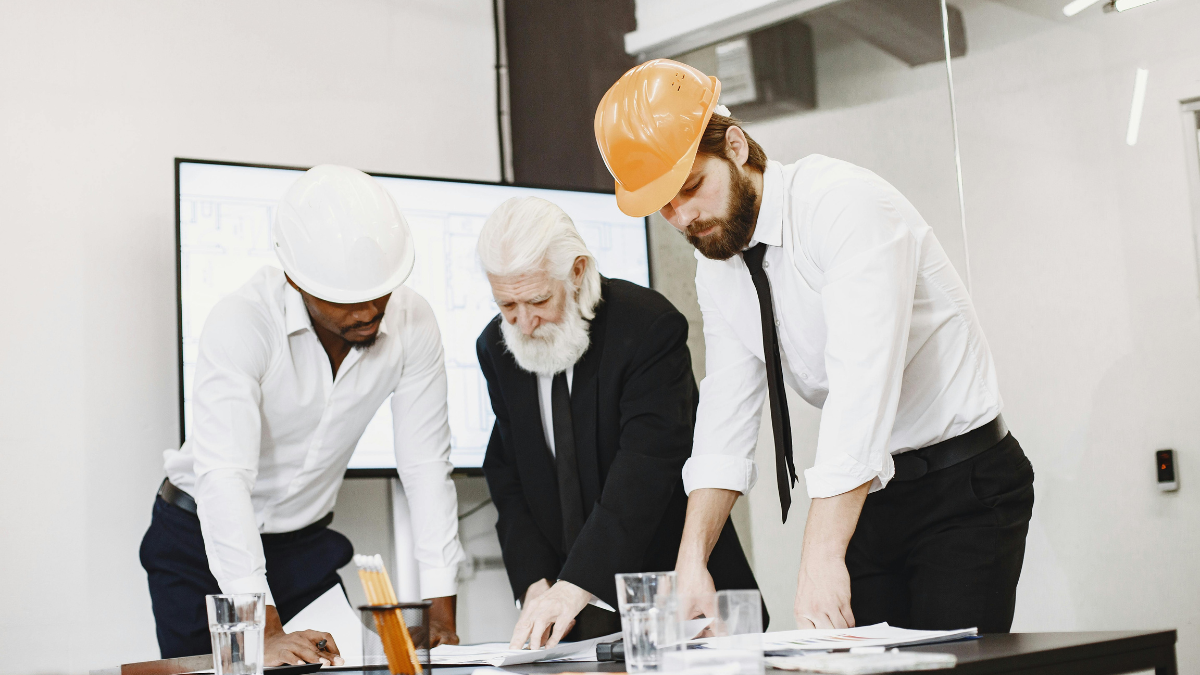 A professional architectural and engineering team collaborates on a complex commercial site plan blueprint outdoors, with a modern commercial building construction visible in the background.
