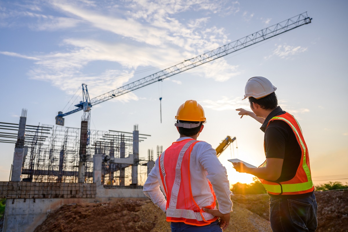 Two construction professionals in safety vests and hard hats review an active building site with a crane at sunset.